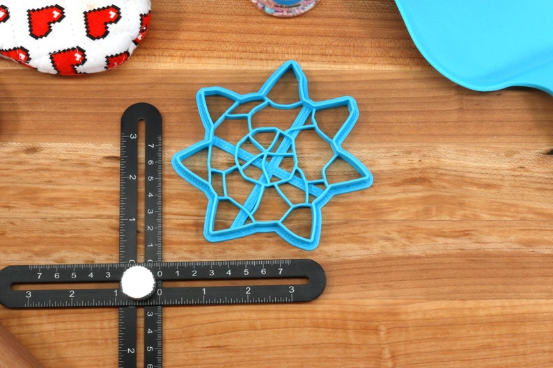 May include: A blue plastic cookie cutter shaped like a star with a geometric design. The cutter is about 3 inches in diameter and is sitting on a wooden surface.