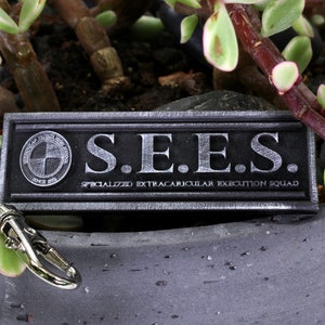 May include: A silver metal keychain with a black rectangular tag that reads "S.E.E.S. SPECIALIZED EXTRACURRICULAR EXECUTION SQUAD".