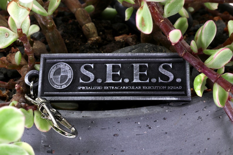 May include: A silver metal keychain with a black rectangular tag that reads "S.E.E.S. SPECIALIZED EXTRACURRICULAR EXECUTION SQUAD".