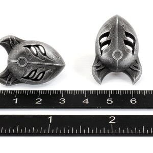 Op de afbeelding: Twee zilverkleurige metalen raketvormige hangers met ingewikkelde ontwerpen. De hangers zijn ongeveer 3,8 cm lang en worden naast een liniaal getoond.
