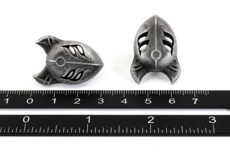 Op de afbeelding: Twee zilverkleurige metalen raketvormige hangers met ingewikkelde ontwerpen. De hangers zijn ongeveer 3,8 cm lang en worden naast een liniaal getoond.