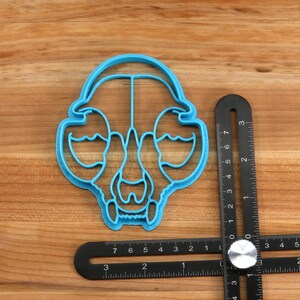 May include: A blue plastic cookie cutter shaped like an animal skull. The cutter is about 2 inches wide and is sitting on a wooden surface next to a black ruler.