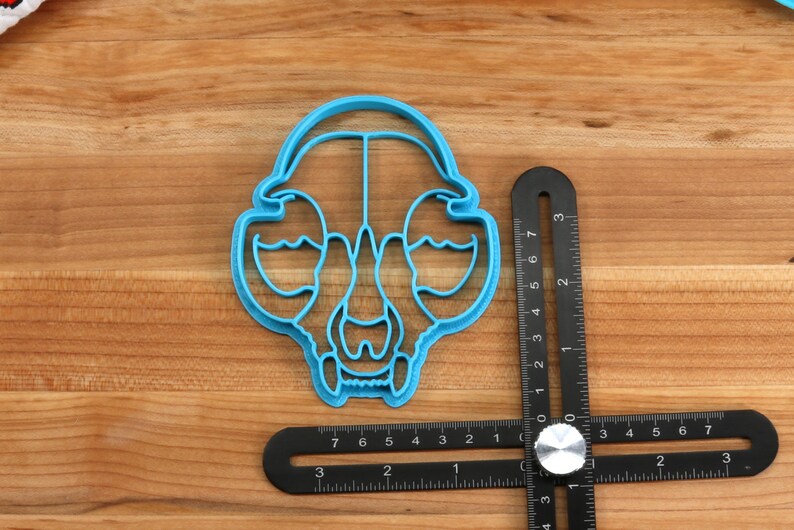 May include: A blue plastic cookie cutter shaped like an animal skull. The cutter is about 2 inches wide and is sitting on a wooden surface next to a black ruler.