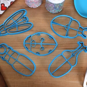 May include: Five blue plastic cookie cutters shaped like different insects, including a ladybug, a beetle, and a dragonfly. The cutters are on a wooden surface.