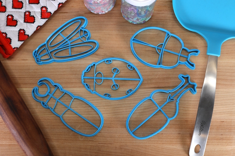 May include: Five blue plastic cookie cutters shaped like different insects, including a ladybug, a beetle, and a dragonfly. The cutters are on a wooden surface.