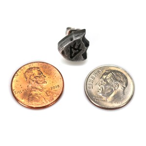 Puede incluir: Un pendiente de metal plateado con un dise&ntilde;o geom&eacute;trico. El pendiente se muestra junto a un centavo estadounidense y una moneda de diez centavos estadounidenses para comparar el tama&ntilde;o.