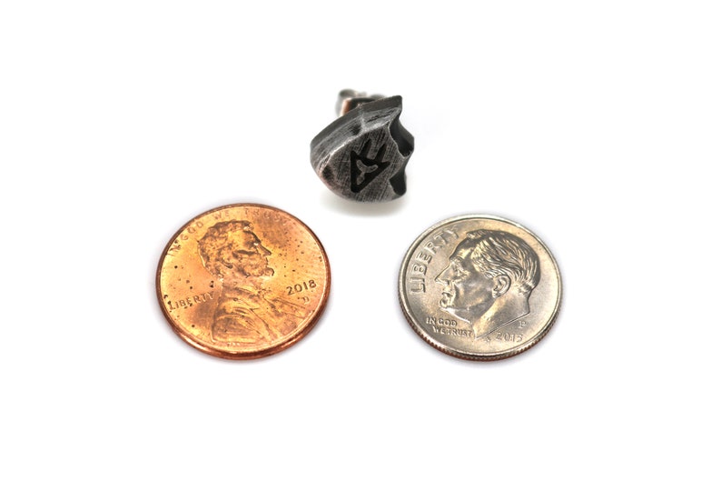 Puede incluir: Un pendiente de metal plateado con un dise&ntilde;o geom&eacute;trico. El pendiente se muestra junto a un centavo estadounidense y una moneda de diez centavos estadounidenses para comparar el tama&ntilde;o.