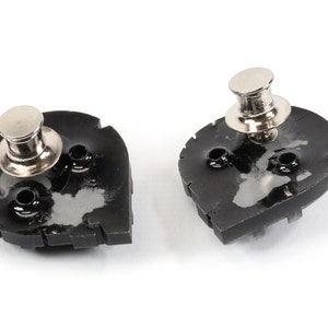 May include: Two black metal snap fasteners with silver metal posts. The fasteners are shaped like a teardrop and have a textured surface.
