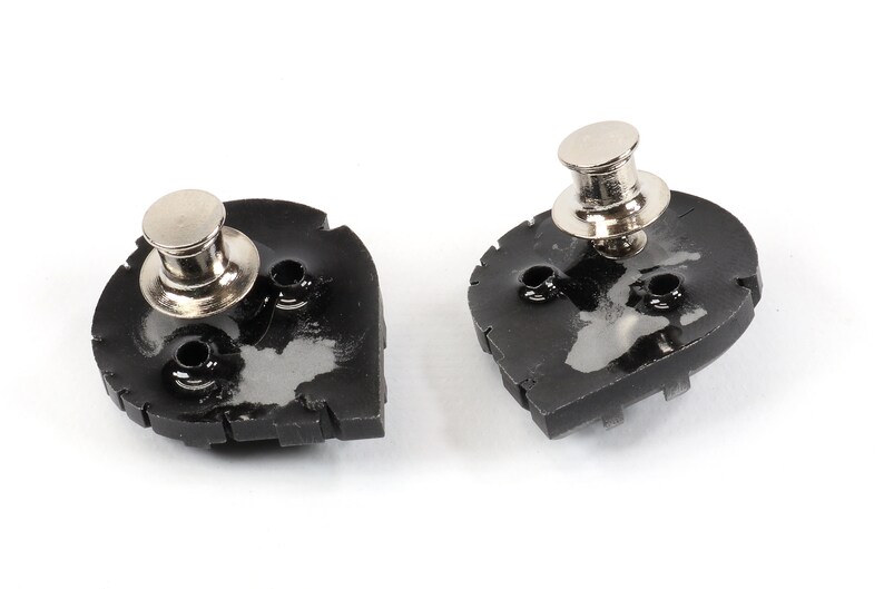 May include: Two black metal snap fasteners with silver metal posts. The fasteners are shaped like a teardrop and have a textured surface.