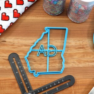 Puede incluir: Un cortador de galletas de pl&aacute;stico azul con forma del estado de Georgia con las letras "AD" en su interior. El cortador est&aacute; sobre una superficie de madera con otros utensilios de reposter&iacute;a.