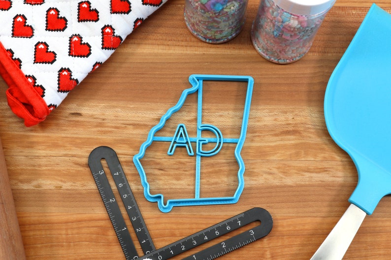 Puede incluir: Un cortador de galletas de pl&aacute;stico azul con forma del estado de Georgia con las letras "AD" en su interior. El cortador est&aacute; sobre una superficie de madera con otros utensilios de reposter&iacute;a.