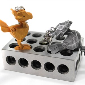 May include: Two 3D printed keychains, one gold and one silver, are displayed on a metal block with multiple holes. The gold keychain is shaped like a bird, while the silver keychain is shaped like a rabbit.