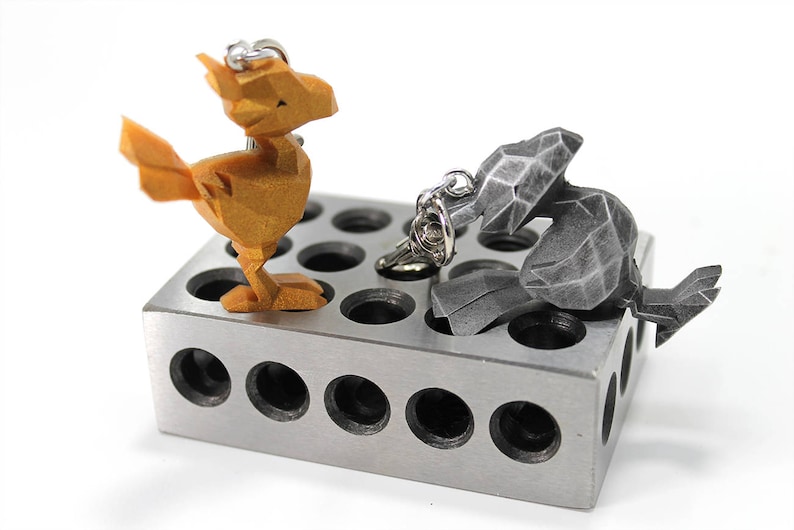 May include: Two 3D printed keychains, one gold and one silver, are displayed on a metal block with multiple holes. The gold keychain is shaped like a bird, while the silver keychain is shaped like a rabbit.