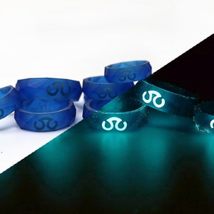 May include: A set of blue silicone rings with a glow-in-the-dark design. The rings are stacked on top of each other and have a geometric pattern. The rings are shown in both daylight and in the dark, highlighting their glow-in-the-dark feature.