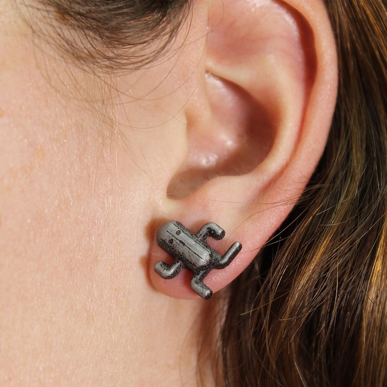 May include: A pair of silver-colored earrings featuring a small, stylized figure with a simple face and limbs.