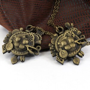 May include: Two gold-toned necklaces with intricate steampunk-style pendants featuring a cartoon-like face with gears and other mechanical details. The necklaces are lying on a brown leather surface.