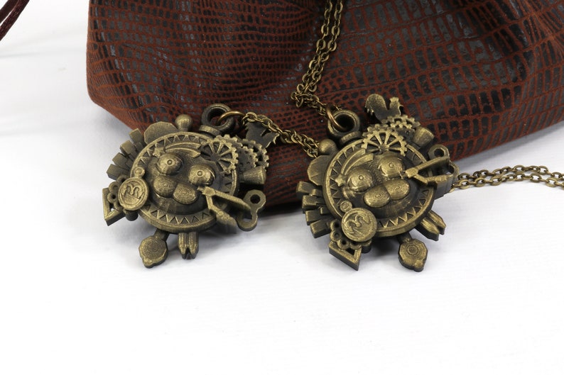 May include: Two gold-toned necklaces with intricate steampunk-style pendants featuring a cartoon-like face with gears and other mechanical details. The necklaces are lying on a brown leather surface.