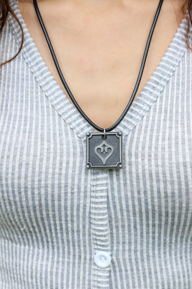 May include: A silver pendant necklace with a square pendant featuring a stylized heart design. The pendant is on a black cord.
