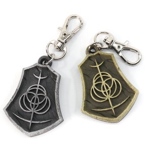 May include: Two keychains with a silver and gold finish. Each keychain features a shield-shaped charm with a stylized symbol of a circle with three smaller circles inside and a vertical line extending from the top.