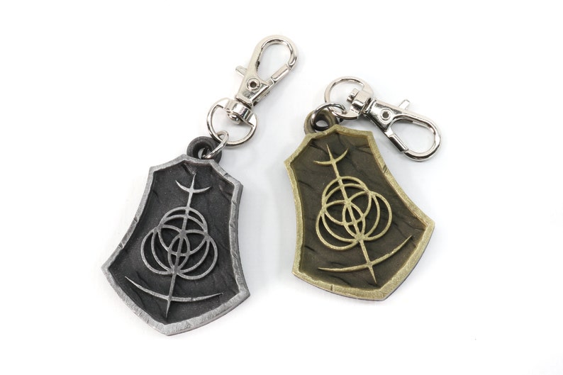 May include: Two keychains with a silver and gold finish. Each keychain features a shield-shaped charm with a stylized symbol of a circle with three smaller circles inside and a vertical line extending from the top.