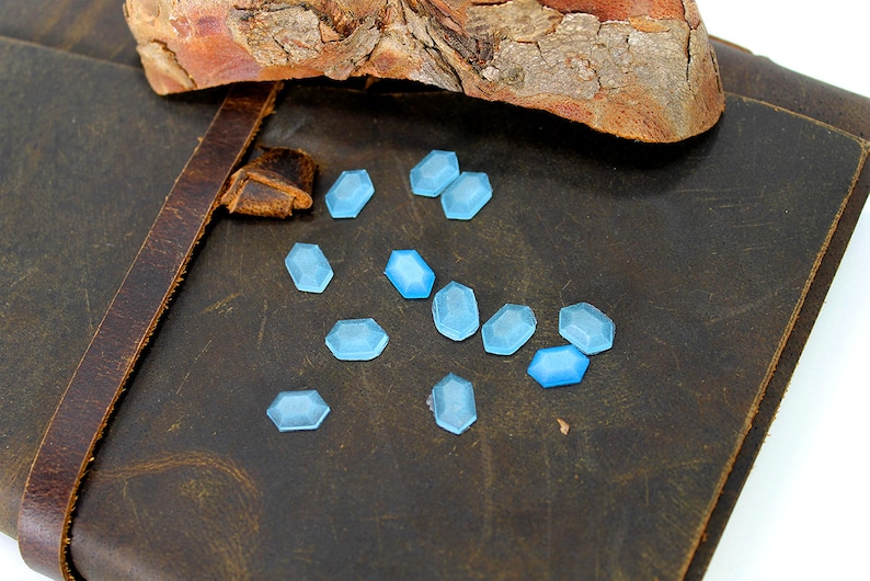 K&ouml;nnte beinhalten: Nahaufnahme eines braunen Lederjournals mit einem Riemen und einem Haufen hellblauer, facettierter Glassteine, die auf der Oberfl&auml;che verstreut sind.