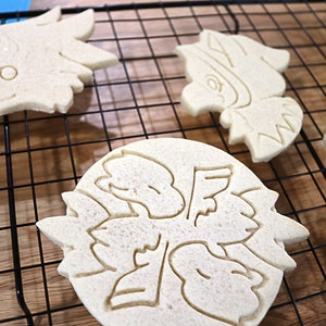 May include: Five plain sugar cookies on a black wire cooling rack. The cookies are in various shapes, including a bird, a flower, and a geometric design.