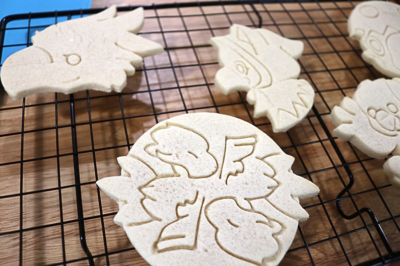 May include: Five plain sugar cookies on a black wire cooling rack. The cookies are in various shapes, including a bird, a flower, and a geometric design.