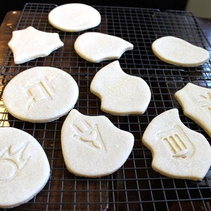 Op de afbeelding: Een close-up van tien witte suikerkoekjes op een draadrek. De koekjes zijn versierd met verschillende symbolen, waaronder een ster, een cirkel met een symbool erin en een schild met een symbool erin.