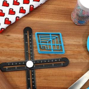 May include: A blue plastic cookie cutter shaped like a geometric design, a black ruler with white markings, a wooden rolling pin, and a blue silicone spatula on a wooden surface.