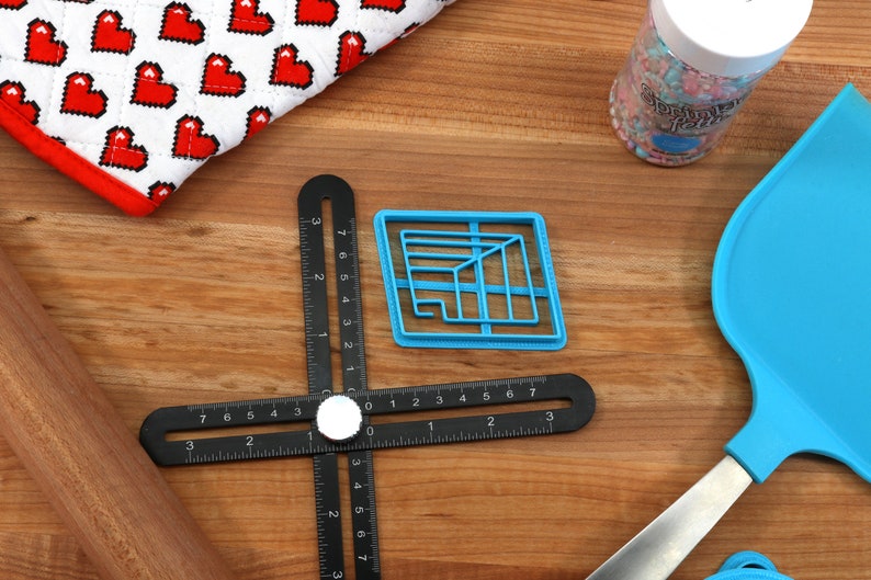 May include: A blue plastic cookie cutter shaped like a geometric design, a black ruler with white markings, a wooden rolling pin, and a blue silicone spatula on a wooden surface.