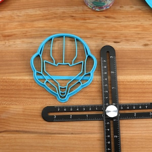 May include: A blue plastic cookie cutter shaped like a video game character's helmet. The cutter is on a wooden surface next to a black ruler.