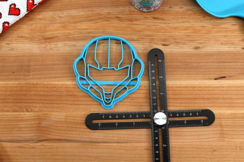 May include: A blue plastic cookie cutter shaped like a video game character's helmet. The cutter is on a wooden surface next to a black ruler.