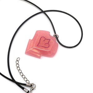 May include: A pink pendant necklace with a black cord. The pendant is a shield shape with a heart design. The necklace has a silver-colored clasp and extender chain. The pendant has a glittery, translucent appearance.