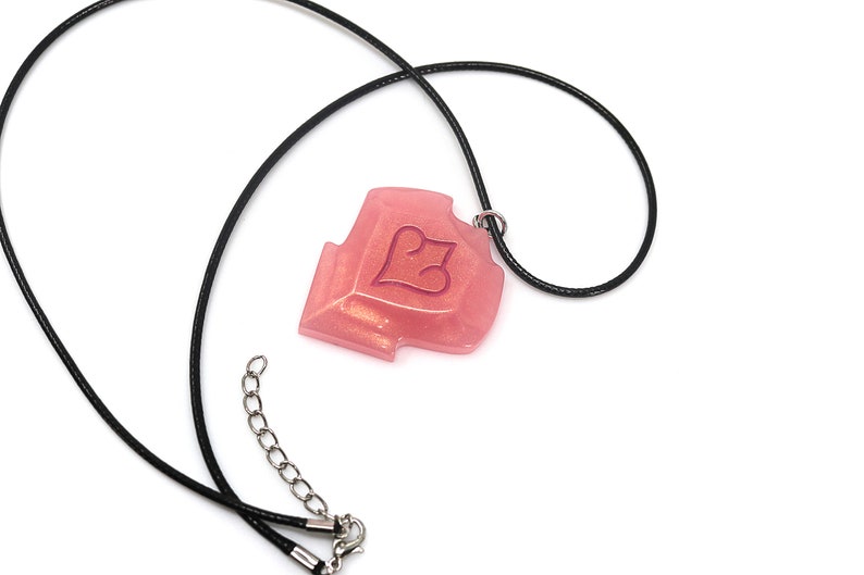 May include: A pink pendant necklace with a black cord. The pendant is a shield shape with a heart design. The necklace has a silver-colored clasp and extender chain. The pendant has a glittery, translucent appearance.