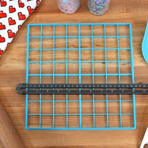 May include: A blue plastic grid with 36 squares, a black ruler, a wooden rolling pin, a blue spatula, and a red and white heart-patterned oven mitt on a wooden surface.