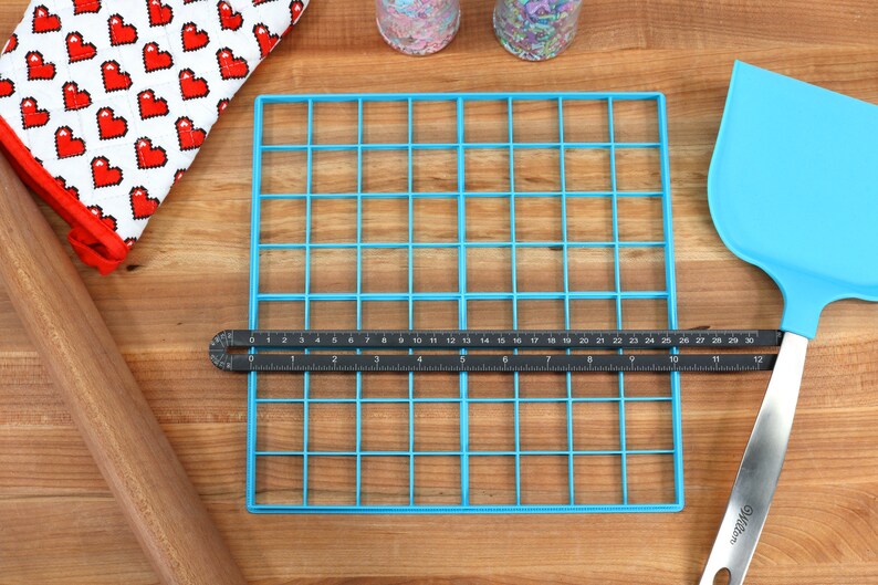 May include: A blue plastic grid with 36 squares, a black ruler, a wooden rolling pin, a blue spatula, and a red and white heart-patterned oven mitt on a wooden surface.