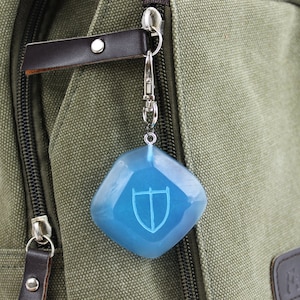 May include: A blue, shield-shaped charm with a white design hangs from a silver keychain attached to a green canvas bag.