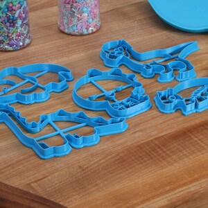 May include: A set of six blue plastic dinosaur cookie cutters on a wooden surface. The cutters are shaped like a stegosaurus, a triceratops, a brachiosaurus, a tyrannosaurus rex, a pterodactyl, and a velociraptor.