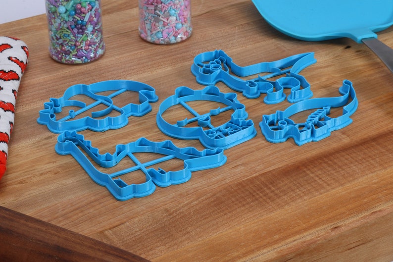 May include: A set of six blue plastic dinosaur cookie cutters on a wooden surface. The cutters are shaped like a stegosaurus, a triceratops, a brachiosaurus, a tyrannosaurus rex, a pterodactyl, and a velociraptor.