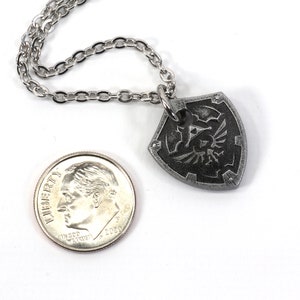 May include: A silver chain necklace with a silver pendant shaped like the Hylian Shield from the Legend of Zelda video game series. The pendant is about the size of a US dime.
