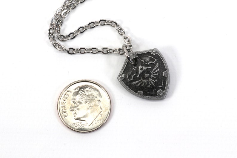 May include: A silver chain necklace with a silver pendant shaped like the Hylian Shield from the Legend of Zelda video game series. The pendant is about the size of a US dime.