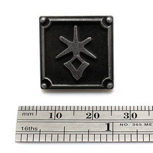 May include: A black metal pin with a stylized design of a star and a diamond shape. The pin is approximately 2 cm wide.