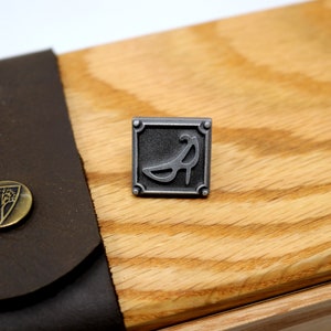 May include: A silver metal pin with a square frame and a stylized image of a high heel shoe inside. The pin is on a wooden surface.