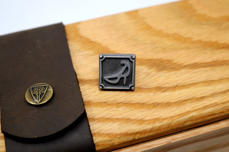 May include: A silver metal pin with a square frame and a stylized image of a high heel shoe inside. The pin is on a wooden surface.