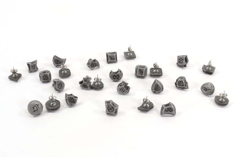 May include: A set of 18 gray earrings with different symbols and designs. The earrings are made of a metal alloy and have a silver finish.
