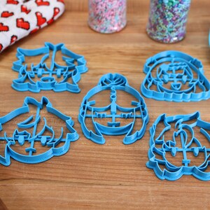 Puede incluir: Cinco cortadores de galletas de pl&aacute;stico azul con forma de caras de dibujos animados. Los cortadores est&aacute;n sobre una superficie de madera.