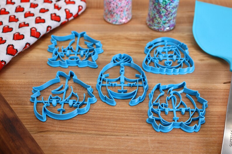 Puede incluir: Cinco cortadores de galletas de pl&aacute;stico azul con forma de caras de dibujos animados. Los cortadores est&aacute;n sobre una superficie de madera.