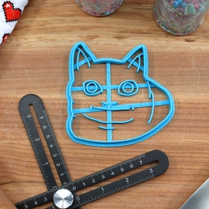 May include: Blue plastic cookie cutter shaped like a cat's face with a wide smile. The cutter has a grid pattern inside the outline of the cat's face.