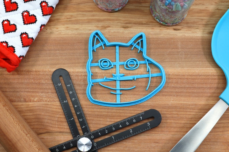 May include: Blue plastic cookie cutter shaped like a cat's face with a wide smile. The cutter has a grid pattern inside the outline of the cat's face.