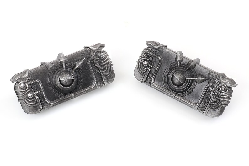 May include: Two silver-coloured metal keychains with intricate designs. Each keychain features a circular centre with a raised arrow and a detailed, textured border.